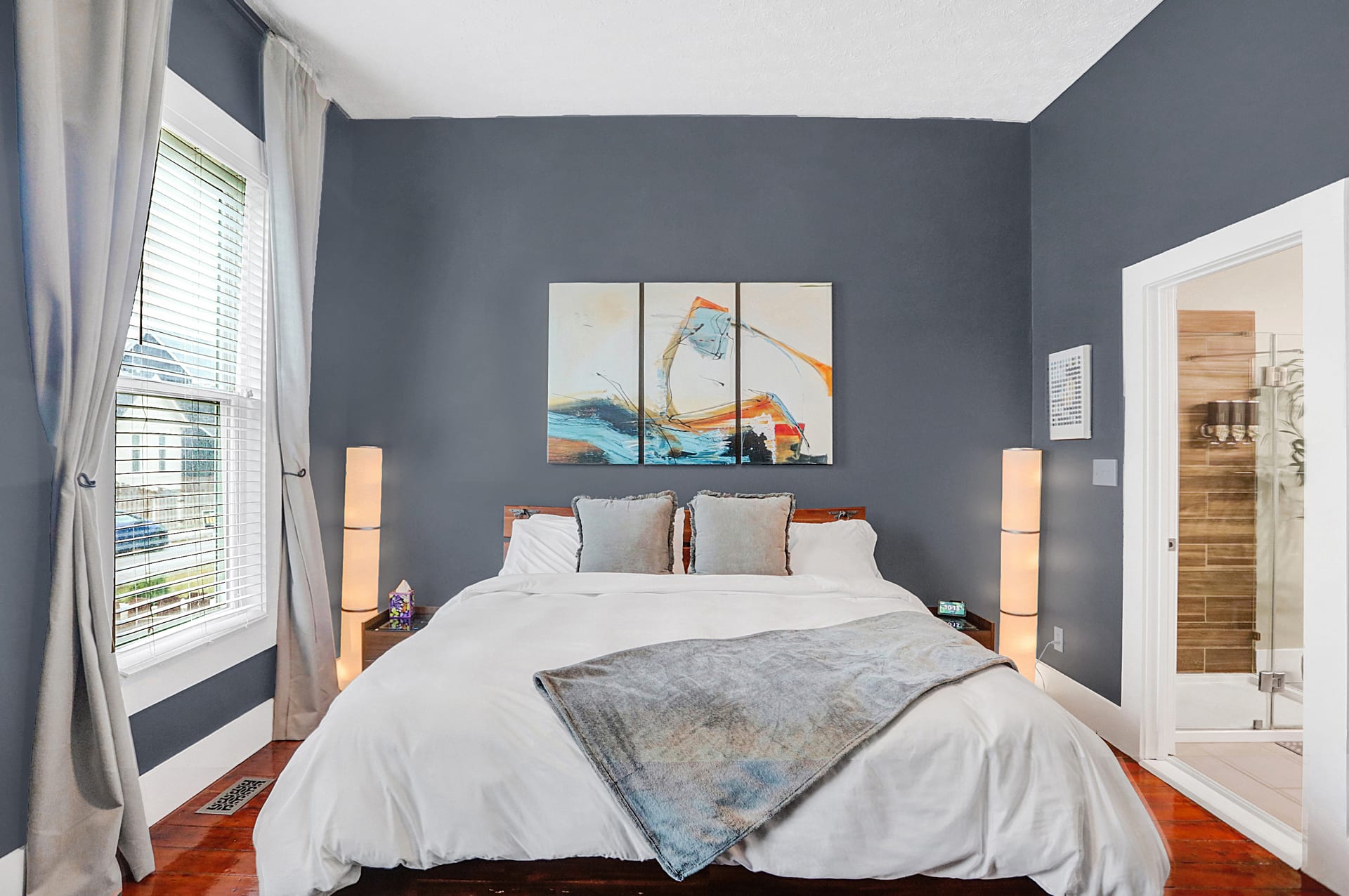 Primary king bedroom with triptych artwork, column lamps, and ensuite bathroom glimpse through the open door