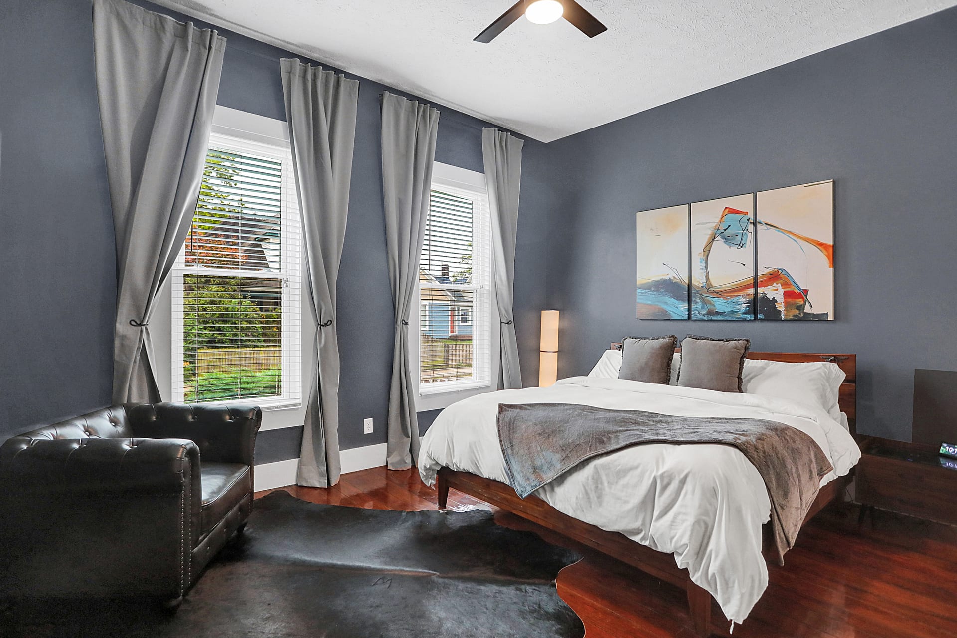 Wide view of the primary bedroom with tall windows, reading chair, cowhide rug, and refinished hardwood floors